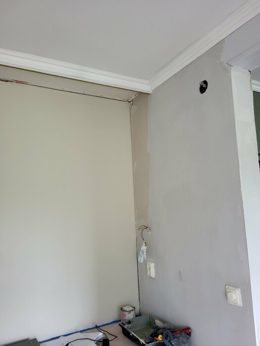 Hörn av rum med borttagna överskåp, synliga elkablar, hål i väggen och målarutrustning på en bänk. Lampknapp och dimmer på höger vägg; målad kabelkanal.