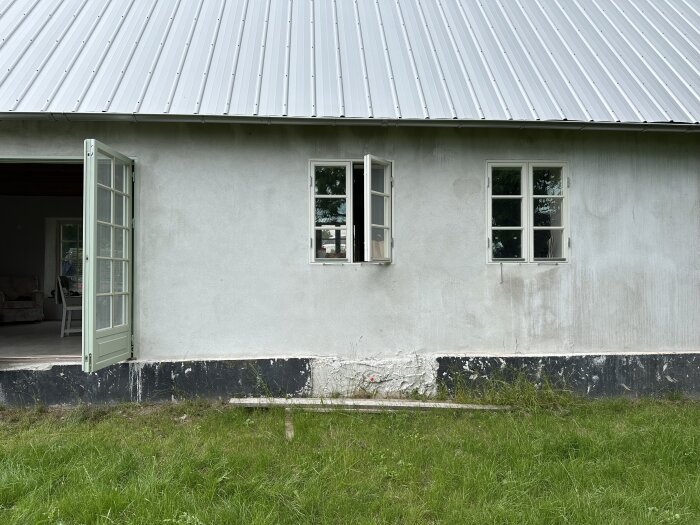 Fasad av hus med vit puts och skadad sockel. Färgsläpp och lagningar syns på sockeln. En grön altandörr och två fönster är öppna. Gräsmatta framför.
