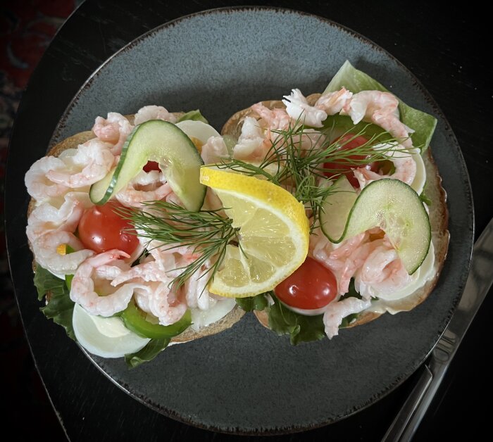 Två räkbaguarnerade smörgåsar med rund botten på en tallrik, toppade med skivor av citron, gurka, tomat och dill.