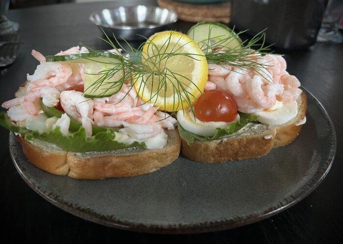 Två räksmörgåsar på en tallrik, toppade med citron, tomat, gurka, ägg, sallad och dill.