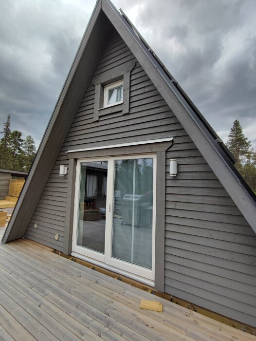 Gråmålat trekantigt hus med liggande träpanel och vita detaljer i fönster och dörr, nymålat med Auson Tjärvitrol. Trädäck i förgrunden och molnig himmel i bakgrunden.