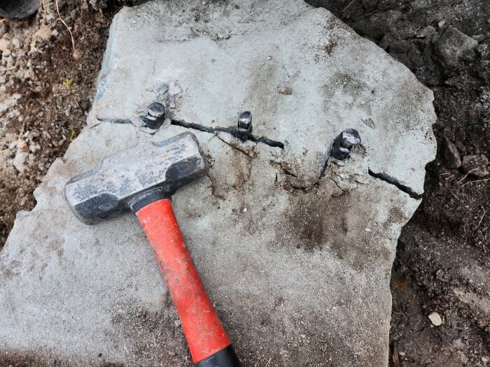 En hammare ligger på en sprucken betongplatta. Fyra bultspetsar sticker upp från plattan. Mark och småsten omger plattan.
