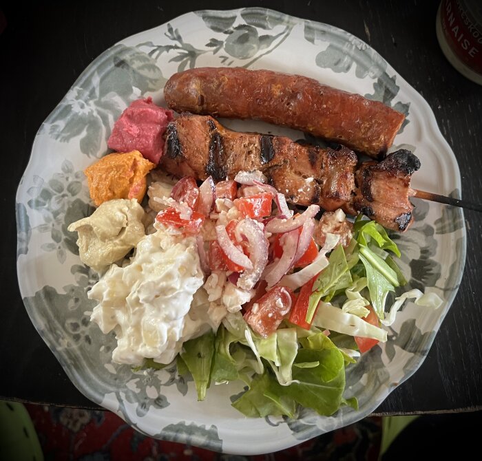 Tallrik med grillad korv, grillspett med griskött, potatissallad, sallad med fetaost och tomat och tre olika sorters hummus: vanlig, rödbeta och chili.