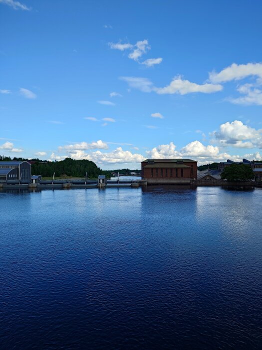 Vy över en sjö med ett rött tegelhus och en damm i bakgrunden under en klarblå himmel med vita moln.