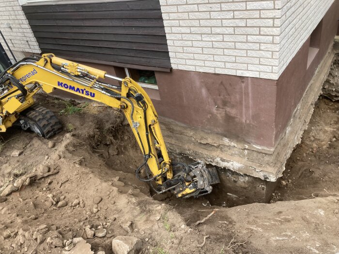 En grävmaskin gräver runt husgrundens kant på ett äldre hus. Husgrunden syns tydligt, delvis utsatt och oisolerad.