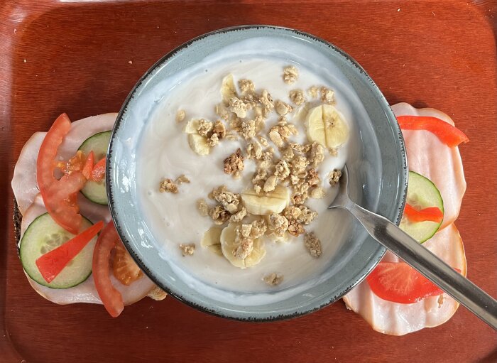 Skål med yoghurt, bananskivor och granola på en bricka med två smörgåsar, en med tomat och gurka, den andra med paprika och gurka, ovanpå en skiva skinka.