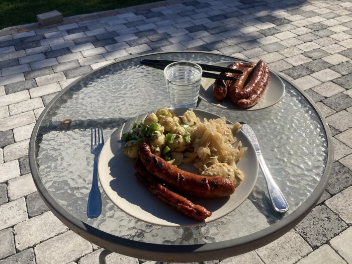 Tallrik med grillad korv, vinegrettpotatissallad och kål på ett glasbord utomhus, med två korvar och ett glas vatten vid sidan.