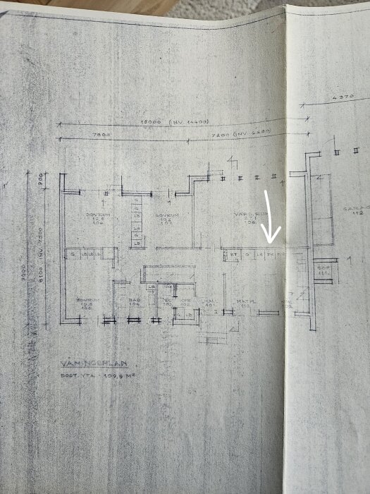 Arkitektritning av ett enplanshus med källare, visar väggar och rumsindelningar; en pil pekar mot väggen mellan köket och vardagsrummet.