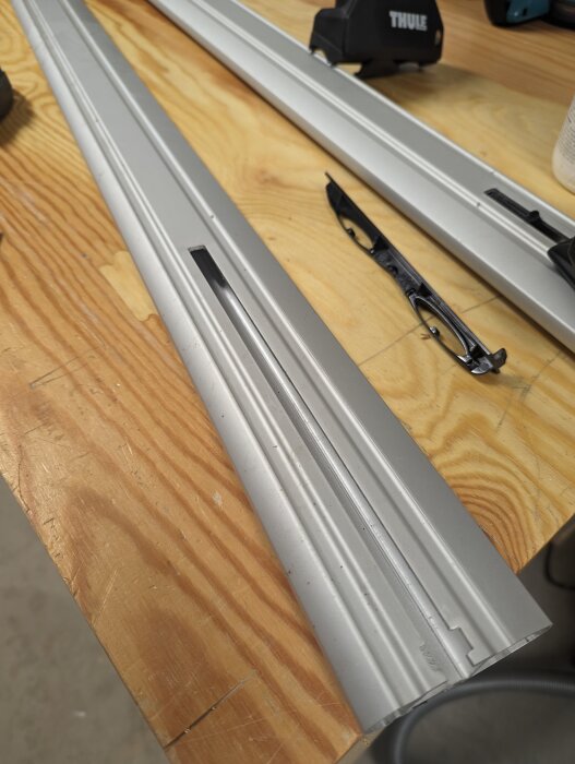 Två aluminiumprofiler med spår ligger på ett arbetsbord av trä, även en svart vinkelslipguider syns på bordet.