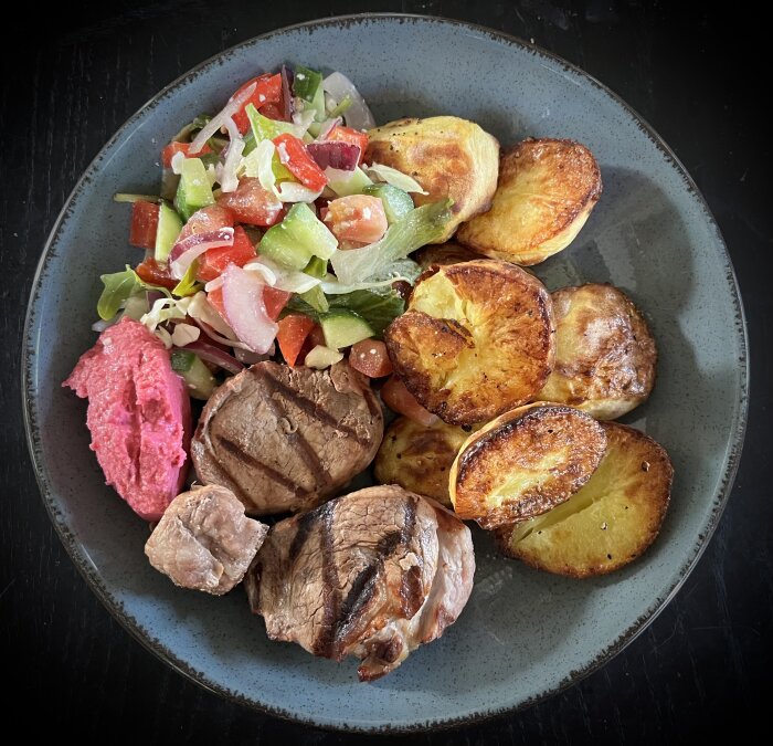 Grillad fläskfilé med ugnsstekt potatis, rödbetshummus och sallad på en tallrik.