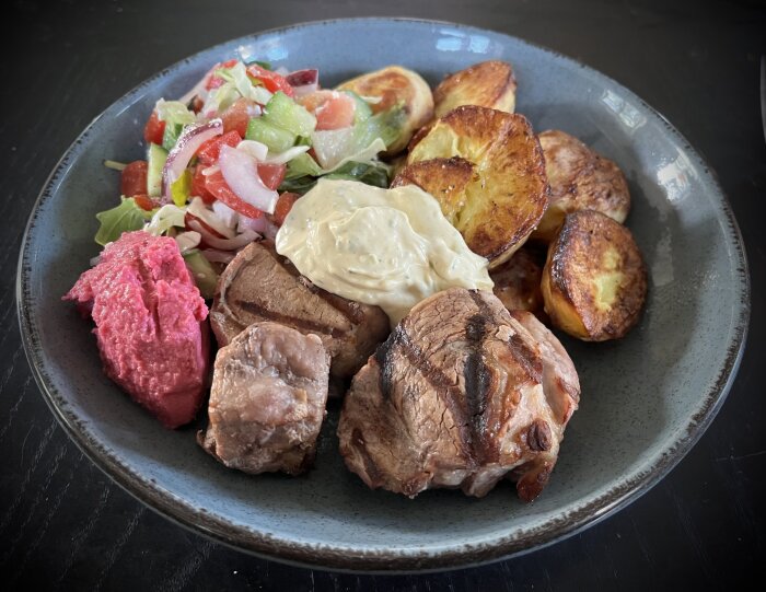 En tallrik med grillad fläskfilé, ugnsstekt potatis, rödbetshummus, sallad med fetaost och tomat samt en klick bearnaisesås.