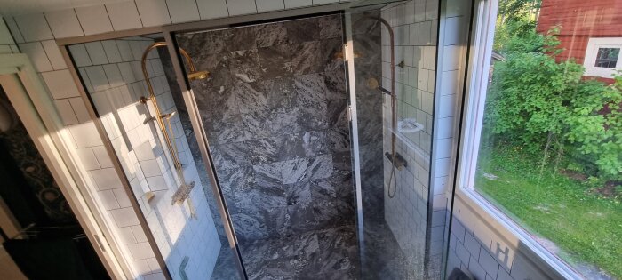 Badrum med svartvitrutiga kakelväggar och dusch med glasdörr, stående bland grönska synlig genom fönstret bredvid duschen.