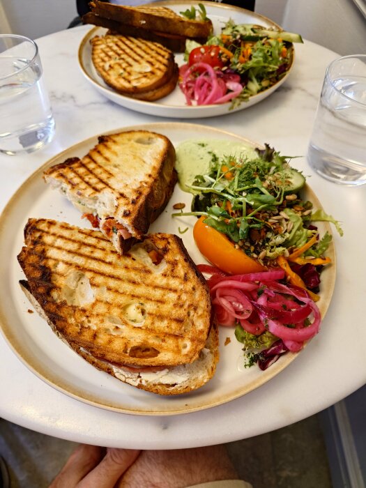 Grillad brie- och salamismörgås på surdegsbröd (levain) med sallad och picklad rödlök på en tallrik, serverad med ett glas vatten på ett bord.