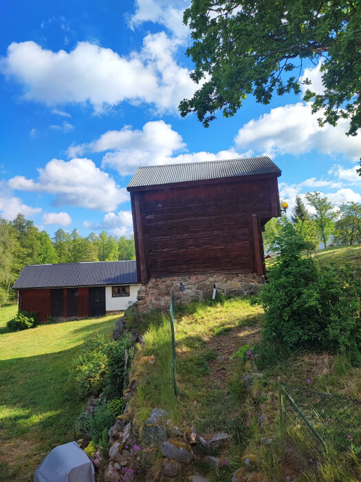 Ett härbre på en sluttning med stenfot, lutande på grund av markförskjutning. En annan byggnad syns i bakgrunden, omgiven av grönska och blå himmel.