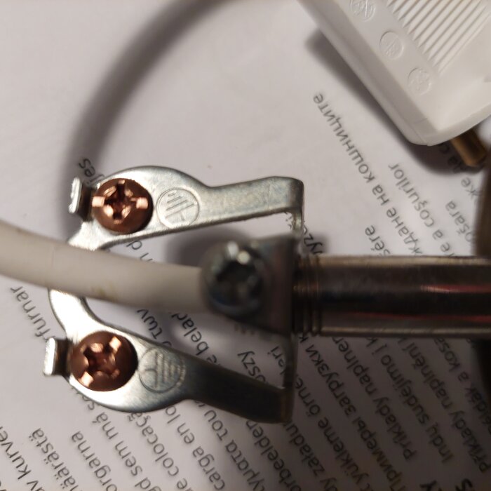 Närbild av en metallklämma som håller en vit kabel på plats, skruvad med en metallskruv och legat på ett papper med text på olika språk.