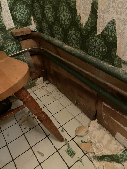 Grön tapet på väggarna i en gästtoalett från 70-talet. Delar av tapeten lossnar och avslöjar fuktskadad trätex bakom. Gamla rör synliga längs nederdelen av väggen.