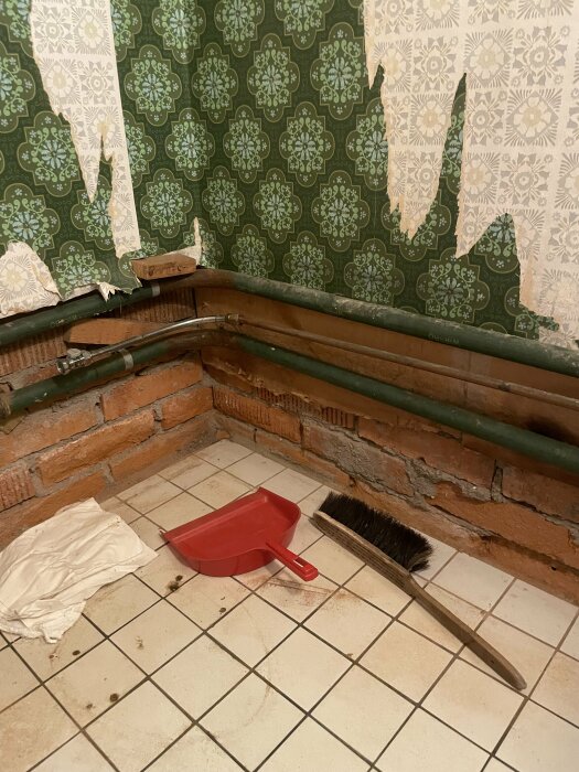 Åldrad gästtoalett med fuktfläckad, avskavd grön tapet och exponerade tegelväggar. Golvet är kaklat och en sopskyffel och kvast ligger på golvet.