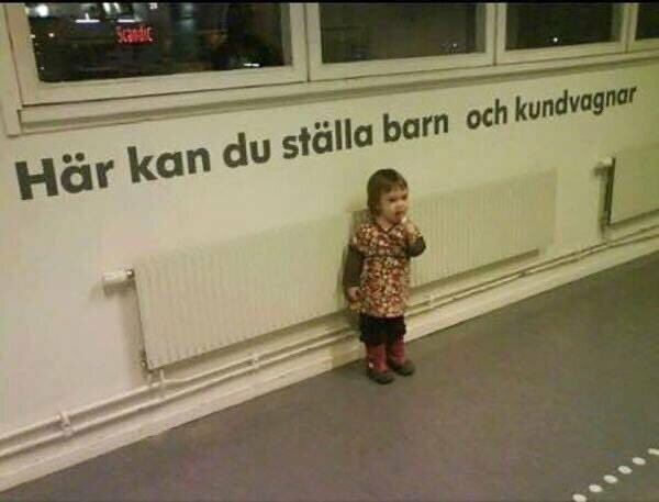 Ett barn står framför en vägg med en skylt som lyder "Här kan du ställa barn och kundvagnar" i en butik eller offentlig plats.