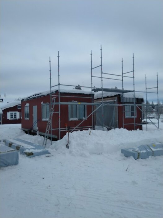 Ett rött fritidshus under konstruktion, täckt av snö på marken och taket, omgivet av byggställningar och byggmaterial.