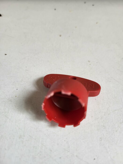 Röd plastdel från en kran på en vit yta,  spetsen är vidgad och har flera taggar runt kanten.