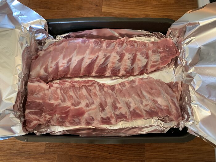 Två råa revbensspjäll ligger i en aluminiumklädd ugnsform, redo att tillagas.