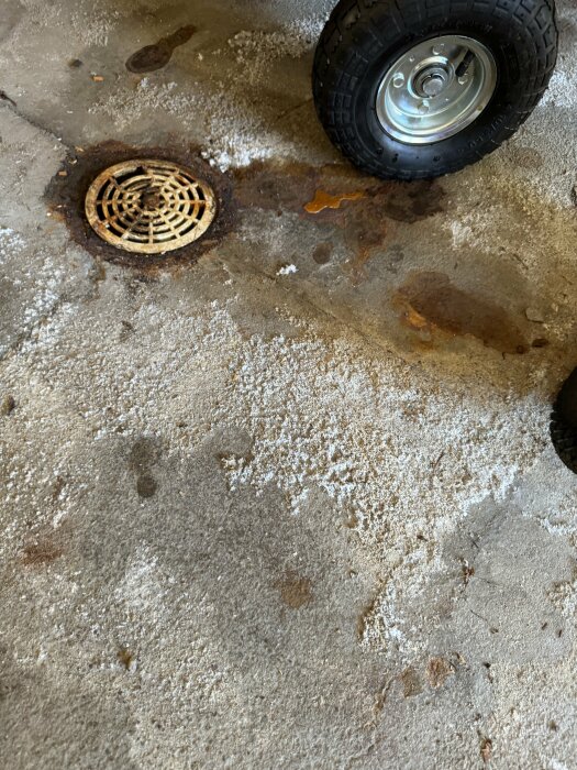 Golvbrunn i garagegolv med rost och smuts kring sargen. Betonggolvet visar saltutfällningar och stillastående vatten nära en hjul.