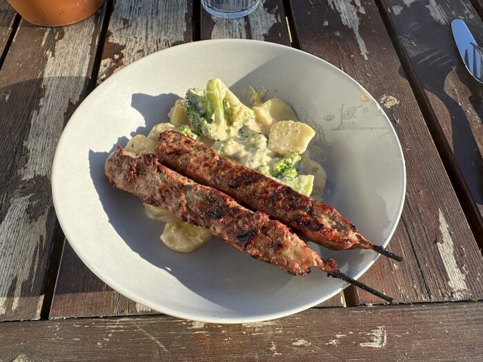 Tallrik med potatisgratäng och två grillspett ovanpå, serverad på ett slitet träbord utomhus i dagsljus.