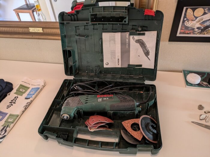 Öppen grön verktygslåda med en Bosch PMF 180 E multiverktyg, flera slip- och sågblad, samt en instruktionsmanual placerad på ett bord.