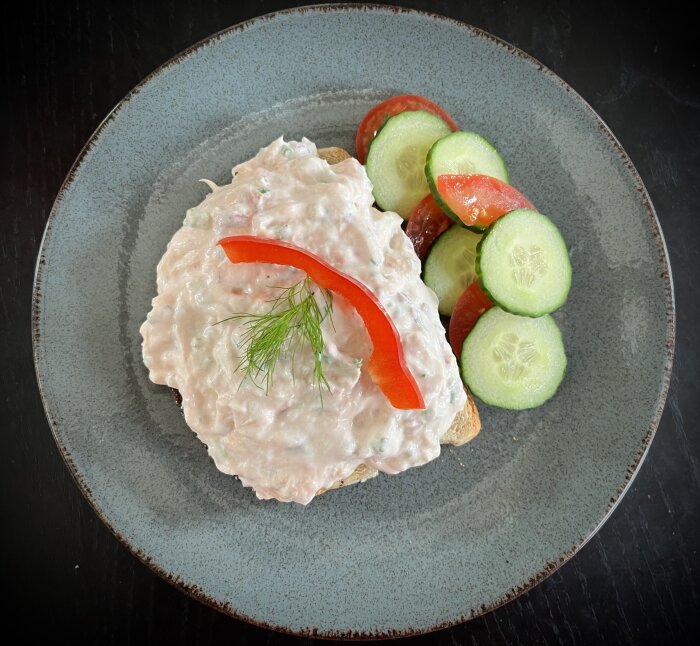 Lax- och räkröra serverad på en rostad brödskiva, toppad med en röd paprikabit och dill, med gurk- och tomatskivor på sidan av tallriken.