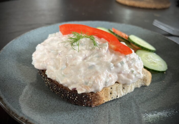 Rostat bröd med laxröra toppad med dill och röd paprika, serverad med skivade gurkor och paprika på en tallrik.