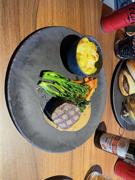 En tallrik med oxfilé, broccoli, morot och gratinerade potatis, serverad på en restaurang med en ölflaska och en burk Coca-Cola Zero.
