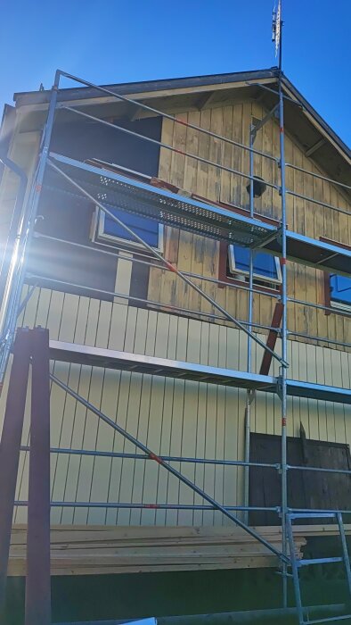 Byggställning rests mot ett hus med delvis borttagen fasad, och den första nya fasadpanelen är monterad.