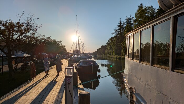 Människor promenerar på en träkaj vid en småbåtshamn i solnedgången med båtar förtöjda vid bryggan.