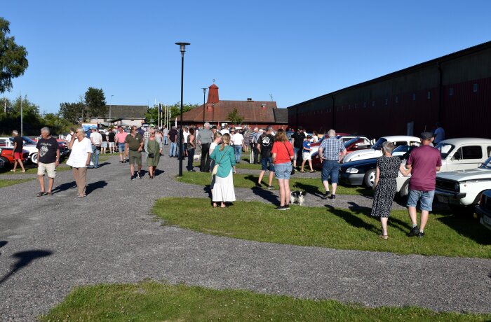 Människor samlade och promenerar bland klassiska bilar under en tisdagsträff i Sunnanå hamn vid Vänern, med blå himmel i bakgrunden.