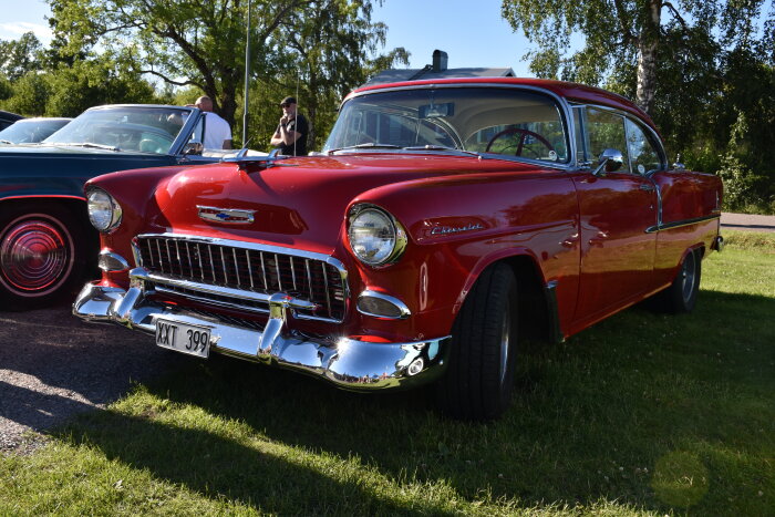 Röd Chevrolet från 1950-talet parkerad på gräset i Sunnanå hamn, omgiven av andra bilar och människor under tisdagskvällen.