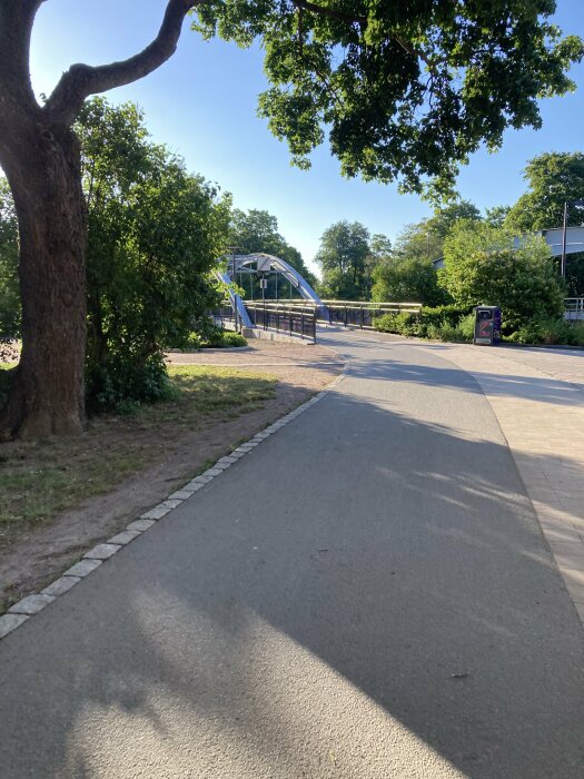 En gångväg genom en park leder till en modern bro i metall. Träd och buskar omger vägen, och solen skiner klart på himlen.