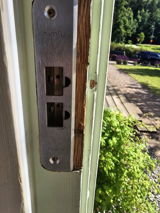Närbild på en dörrkarm med en låsplatta där texten "nimly" syns, omgiven av trädgårdsmiljö i bakgrunden.