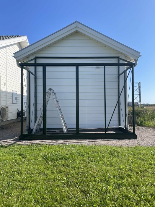 Vägghängt växthus monterat mot en vit träpanelvägg på ett carportförråd under uppbyggnad, stege placerad inuti stommen.