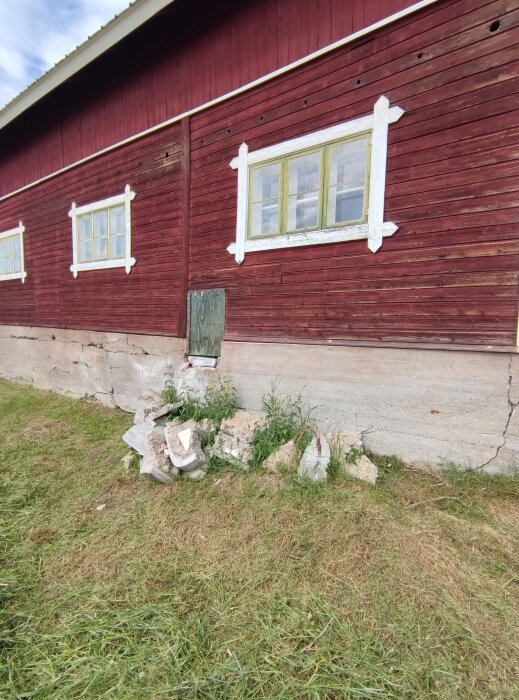 Röd ladugård med vitmålade fönsterkarmar och sprucken, förskjuten grund; marken vid grunden visar spruckna betongdelar och ogräs.