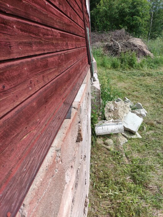 En sprucken grund av betong och en röd trävägg på en gammal ladugård från 1940-talet, med bitar av sten och trädgrenar i gräset vid sidan av byggnaden.