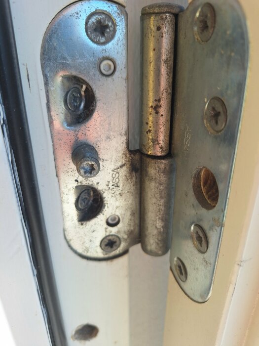 Närbild på ett silverfärgat dörrgångjärn monterat på en vit dörrkarm. Gångjärnet visar slitage och rost, samt flera skruvar med slitna skruvhuvuden.
