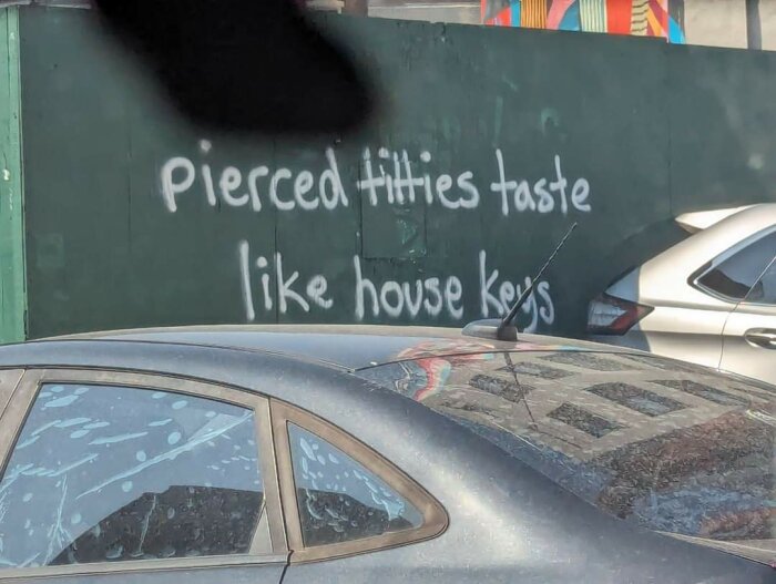 Graffiti-text på en grön vägg som lyder: "Pierced titties taste like house keys." Framför väggen står två bilar parkerade.