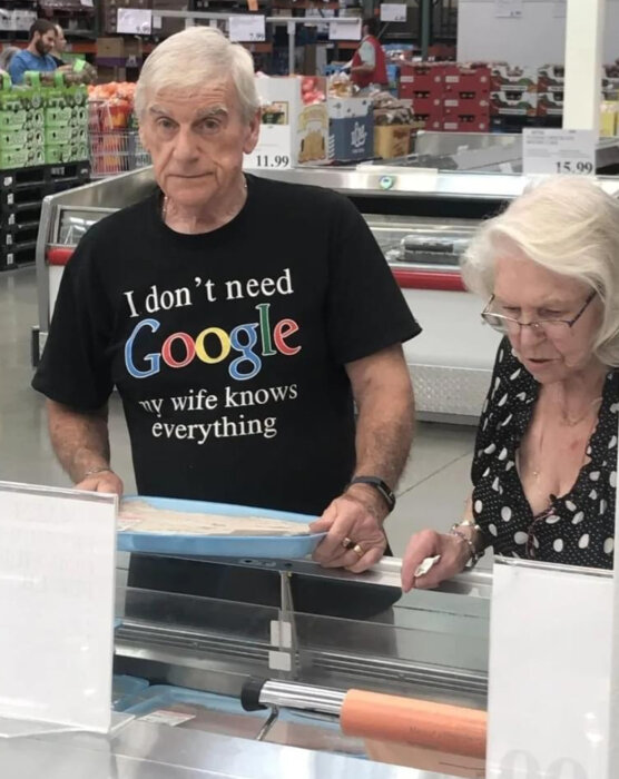 Två äldre personer handlar i en mataffär, mannen bär en t-shirt med texten "I don't need Google, my wife knows everything".