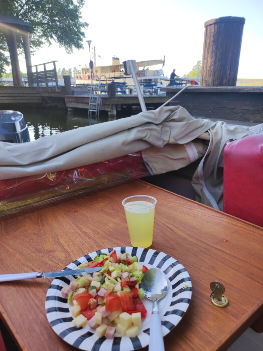 En tallrik med sallad bredvid ett glas saft på ett träbord vid vatten, med en båt och brygga i bakgrunden.