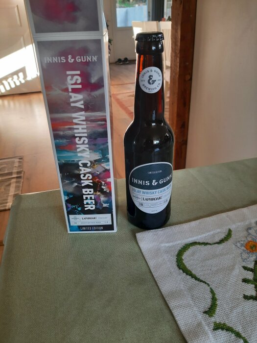 En flaska med etiketten "Innis & Gunn Islay Whisky Cask Beer" och dess förpackning på ett bord i ett rum med en matta och bordsduk.