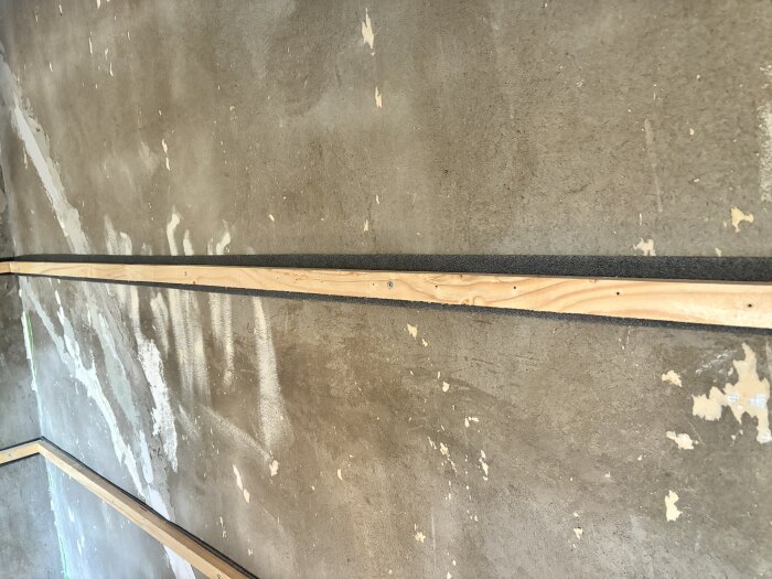 Träreglar med syllisolering monterade mot en betongvägg i en byggprocess. Betongväggen har synliga spår av tidigare cellplastisolering.