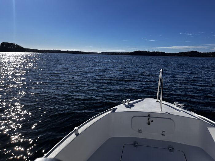 Båt förtöjd på sjö med solglittrande vatten och trädkantad horisont under en klarblå himmel.