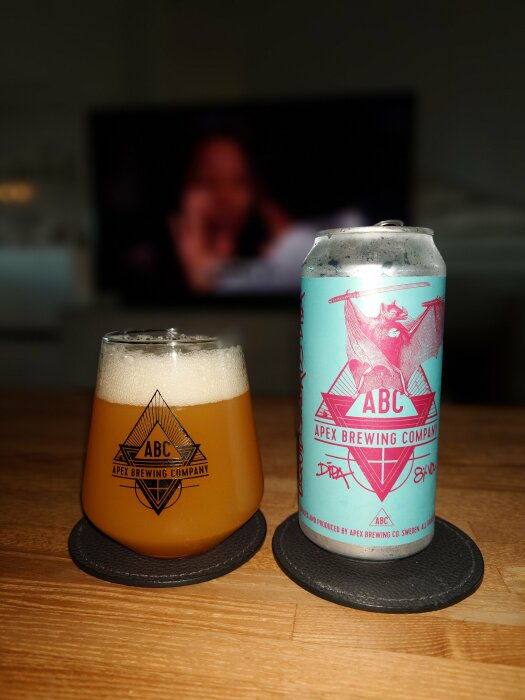 En burk och ett glas Apex öl ståendes på ett bord med en TV i bakgrunden. Burkens design är blå med Apex Brewing Companys logga och en illustration av en fladdermus.