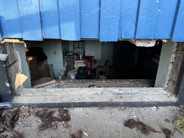 Ovalt hål i grund under blå fasad där fönster tidigare suttit, synligt inåt källare med ojämnt och smutsigt underlag, verktyg och material i bakgrunden.