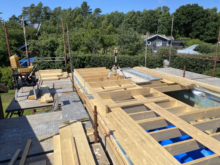 Byggarbetsplats med träbjälklag och planlaser på stativ för att kontrollera nivåer. Solsken, omgivande träd och hus i bakgrunden.